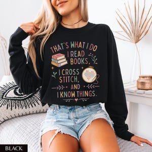May include: Black sweatshirt with the text "THAT'S WHAT I DO I READ BOOKS, I CROSS STITCH, AND I KNOW THINGS." The graphic includes books and a cross-stitch design. The sweatshirt is worn with denim shorts.