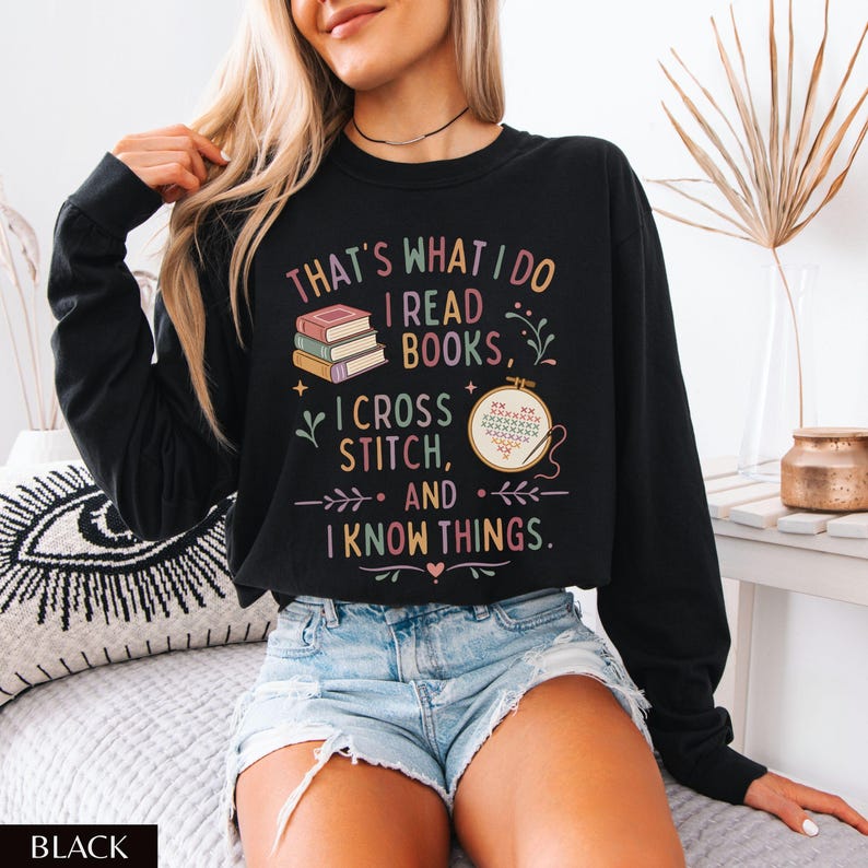 May include: Black sweatshirt with the text "THAT'S WHAT I DO I READ BOOKS, I CROSS STITCH, AND I KNOW THINGS." The graphic includes books and a cross-stitch design. The sweatshirt is worn with denim shorts.