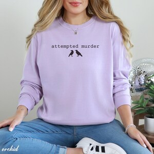 May include: A light purple long-sleeve sweatshirt with the text "attempted murder" above two black bird silhouettes. The sweatshirt is made of a soft material and is a comfortable fit.