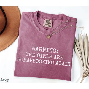 May include: A berry-colored t-shirt with the text "WARNING: THE GIRLS ARE SCRAPBOOKING AGAIN" in white. A gold necklace and a brown hat are also visible. The shirt has a crew neck and a Comfort Colors tag.