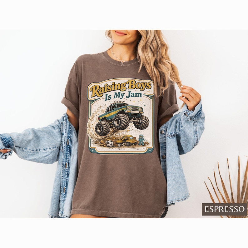 May include: Espresso-colored t-shirt with a graphic of a monster truck jumping over a soccer ball and a small car. The text "Raising Boys Is My Jam" is displayed in a vintage style. The shirt is worn by a person.