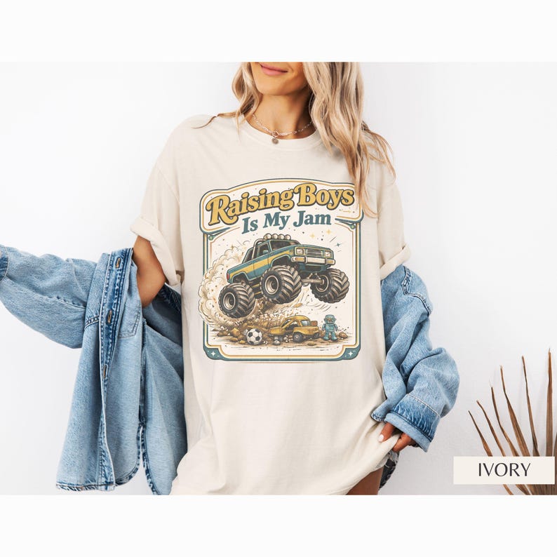 May include: Ivory-colored t-shirt featuring a graphic of a monster truck jumping over a small car, with the text "Raising Boys Is My Jam" in a vintage style. The shirt is worn by a person with blonde hair, wearing a denim jacket.