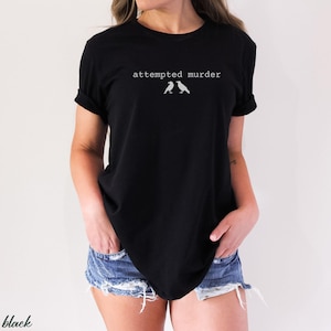 Pu&ograve; includere: T-shirt nero con la scritta "attempted murder" sopra due piccole illustrazioni di uccelli. La maglietta &egrave; a girocollo e viene indossata con pantaloncini di jeans.