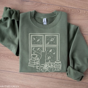 May include: Military green sweatshirt with a white graphic of a cat looking out a window at falling leaves. The window scene includes a stack of books, a cup of coffee, pumpkins, a candle, and a cookie jar.
