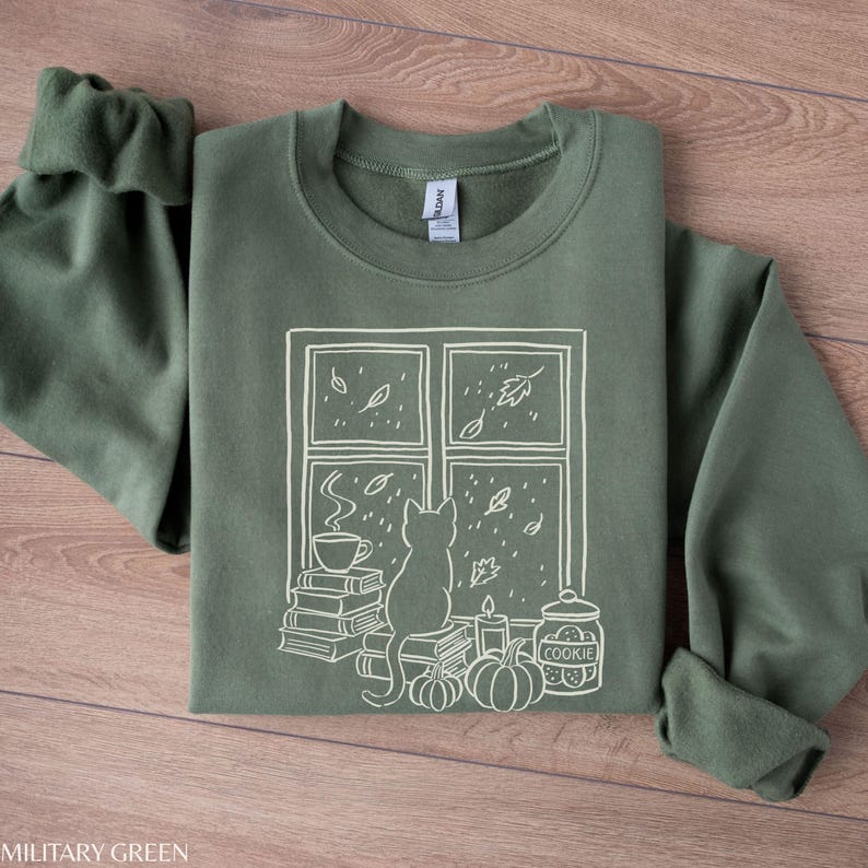 May include: Military green sweatshirt with a white graphic of a cat looking out a window at falling leaves. The window scene includes a stack of books, a cup of coffee, pumpkins, a candle, and a cookie jar.
