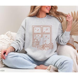 May include: A light gray sweatshirt with a brown graphic of a cat looking out a window at falling leaves. The window scene includes a stack of books, a cup of coffee, pumpkins, and a cookie jar. The word "COOKIE" is visible on the jar.