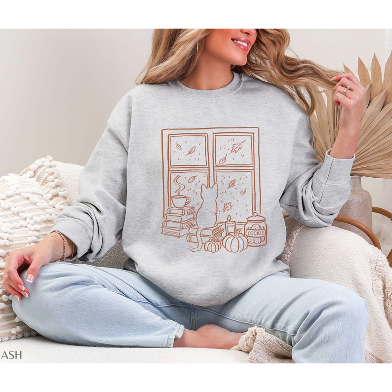 May include: A light gray sweatshirt with a brown graphic of a cat looking out a window at falling leaves. The window scene includes a stack of books, a cup of coffee, pumpkins, and a cookie jar. The word "COOKIE" is visible on the jar.