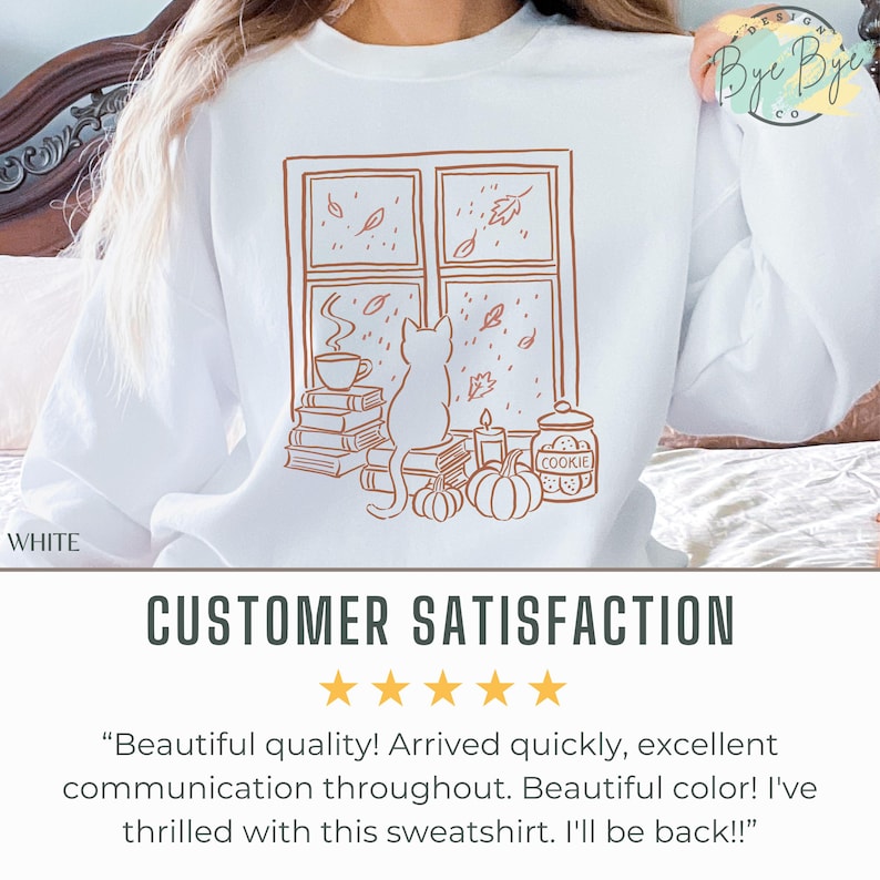 May include: White sweatshirt featuring a brown line drawing of a cat looking out a window at falling leaves. A cup of coffee, books, pumpkins, a candle, and a cookie jar are also depicted. The word "WHITE" is printed on the bottom left.