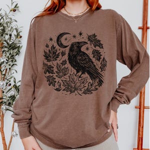 May include: A brown long-sleeve crewneck sweatshirt featuring a black graphic print on the chest. The design depicts a raven perched among various autumn leaves, stars, and a crescent moon, all arranged in a circular composition.