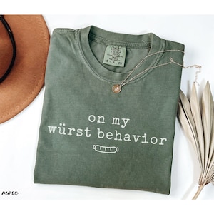 May include: A moss green Comfort Colors t-shirt with the phrase "on my würst behavior" in white text, along with a sausage graphic. A gold necklace and a brown hat are also visible.