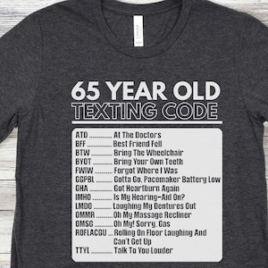 May include: A dark gray t-shirt with white text that reads "65 Year Old Texting Code" and a list of abbreviations and their meanings. The abbreviations are common texting slang, such as ATD for "At The Doctors" and BFF for "Best Friend Fell".