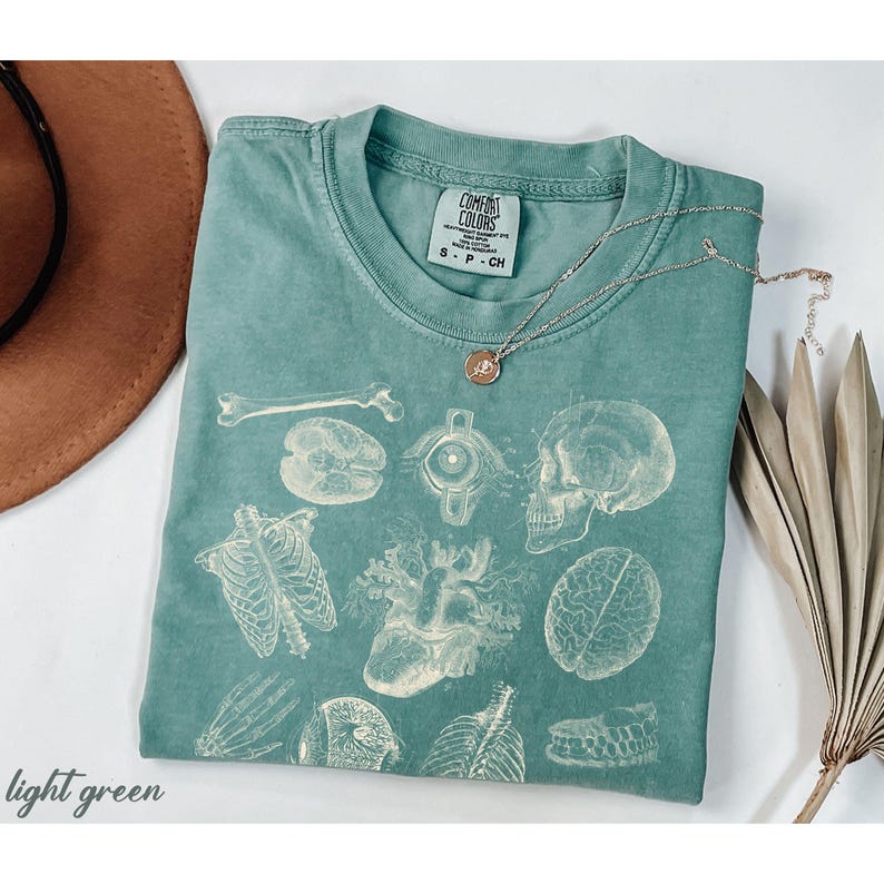 May include: Light green Comfort Colors t-shirt with a vintage anatomical print of bones, organs, and a skull. A gold necklace and a brown hat are next to the shirt. The words "light green" are at the bottom left.