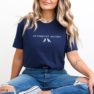 Pu&ograve; includere: T-shirt blu navy con la scritta "attempted murder" e due sagome di uccelli. La maglia ha un girocollo e maniche corte. La modella indossa jeans blu.