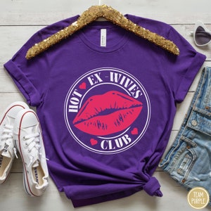 May include: Purple t-shirt with a white circle graphic that says "Hot Ex-Wives Club" with a red lip print inside the circle and red hearts around the circle.
