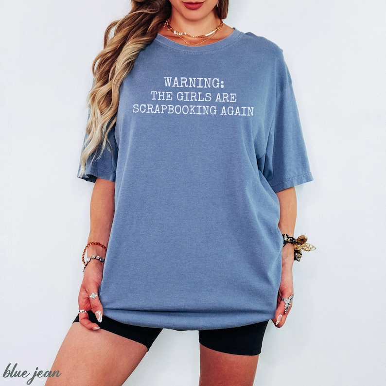 May include: A blue jean-colored t-shirt with the text "WARNING: THE GIRLS ARE SCRAPBOOKING AGAIN" in white. The shirt is a relaxed fit and has short sleeves. The model is wearing black shorts and jewelry.