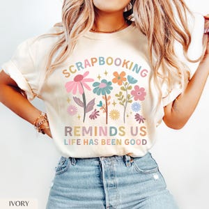 May include: An ivory t-shirt featuring a colorful graphic of various flowers and the text SCRAPBOOKING REMINDS US LIFE HAS BEEN GOOD. The design includes pink, blue, orange, and yellow floral illustrations with small star accents.