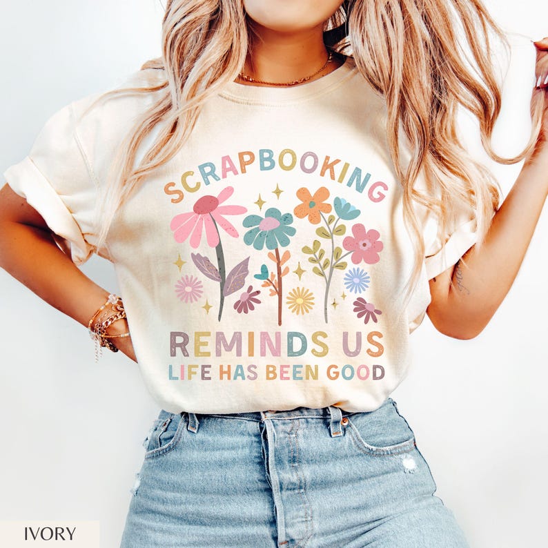 May include: An ivory t-shirt featuring a colorful graphic of various flowers and the text SCRAPBOOKING REMINDS US LIFE HAS BEEN GOOD. The design includes pink, blue, orange, and yellow floral illustrations with small star accents.
