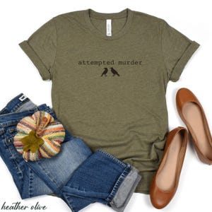 Pu&ograve; includere: T-shirt verde oliva melange con la scritta "attempted murder" e due sagome di uccelli neri. La maglietta &egrave; abbinata a jeans blu, una zucca decorativa e ballerine marroni.