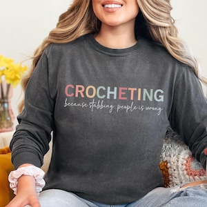 May include: A dark gray long-sleeved sweatshirt with the text "CROCHETING because stabbing people is wrong" in rainbow colors.