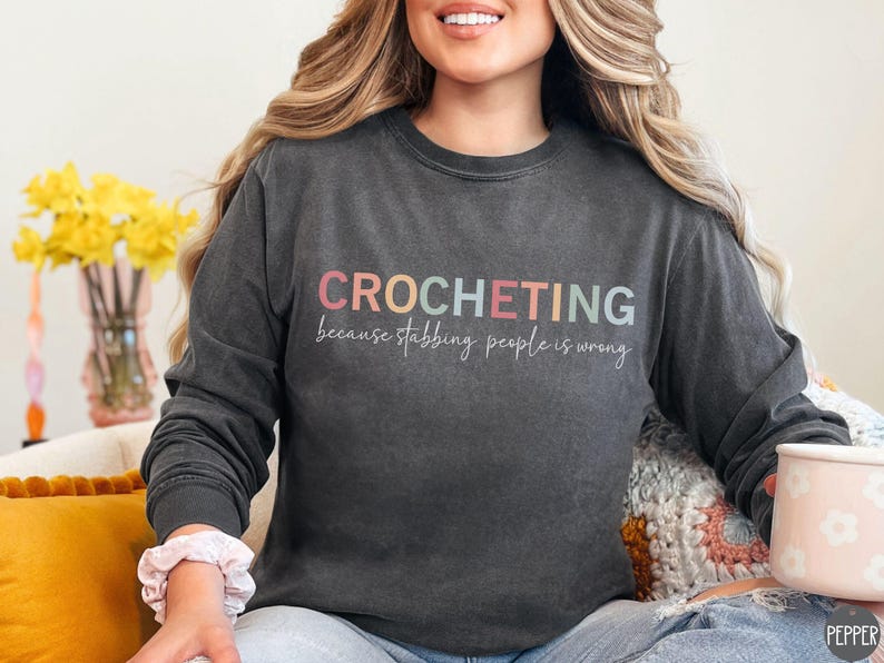 May include: A dark gray long-sleeved sweatshirt with the text "CROCHETING because stabbing people is wrong" in rainbow colors.