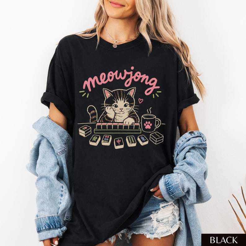 May include: A black t-shirt featuring a graphic of a cat playing mahjong with the text meowjong in pink letters. The design includes mahjong tiles, a mug with a paw print, and a small heart. The shirt has short sleeves and a crew neckline.
