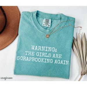 May include: Seafoam green t-shirt with the text "WARNING: THE GIRLS ARE SCRAPBOOKING AGAIN" in white. A gold necklace with a pendant and a brown hat are next to the shirt. The shirt is labeled "COMFORT COLORS".
