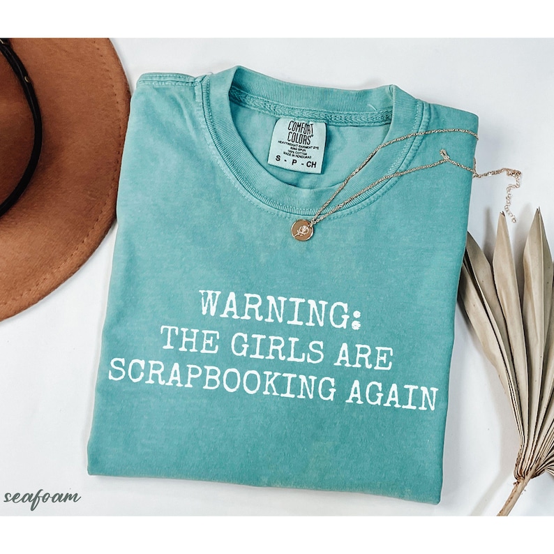May include: Seafoam green t-shirt with the text "WARNING: THE GIRLS ARE SCRAPBOOKING AGAIN" in white. A gold necklace with a pendant and a brown hat are next to the shirt. The shirt is labeled "COMFORT COLORS".