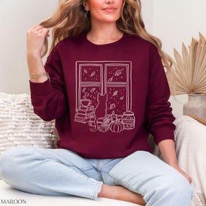 May include: Maroon sweatshirt with a white graphic of a cat sitting by a window, with books, pumpkins, and a cookie jar. The window shows falling leaves and a cup of coffee. The word "MAROON" is printed at the bottom.