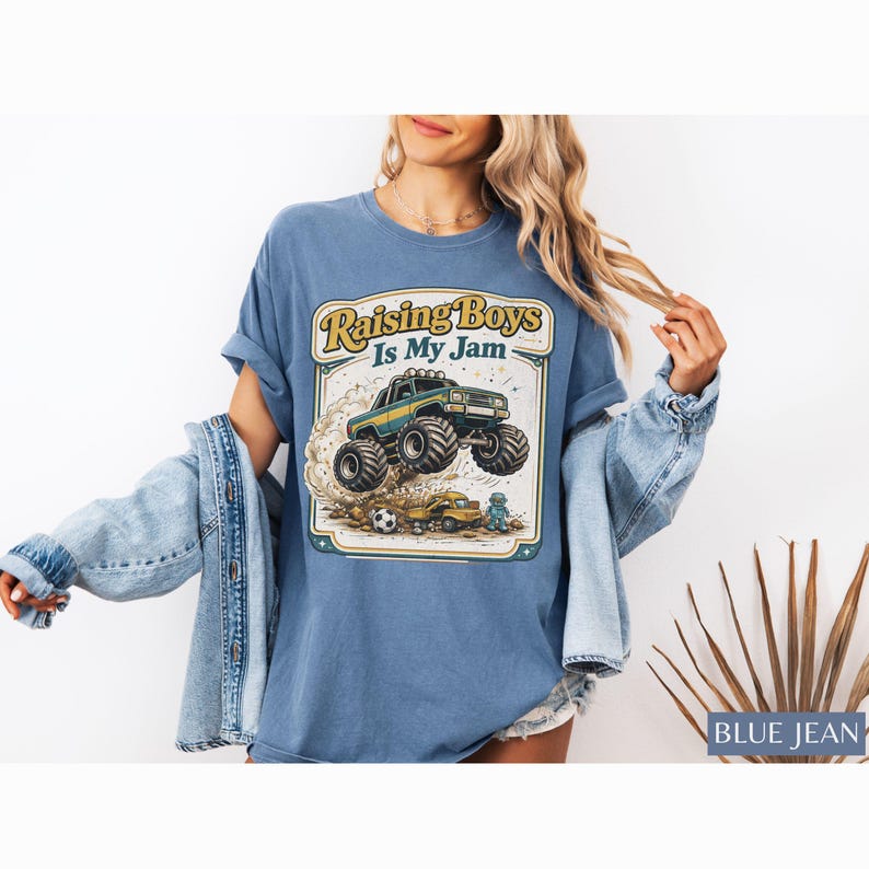 May include: Blue jean t-shirt with a graphic that says "Raising Boys Is My Jam." The graphic features a monster truck, a soccer ball, and a small toy truck. The shirt is a light blue color.