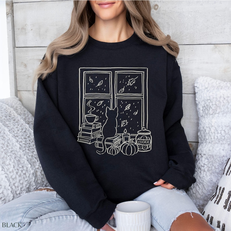 May include: Black sweatshirt with a white graphic of a cat looking out a window at falling leaves. The window scene includes a stack of books, a cup of coffee, pumpkins, a candle, and a cookie jar. The word "BLACK" is printed at the bottom left.
