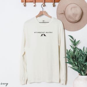 May include: Ivory long-sleeve shirt with the text "attempted murder" above two black bird silhouettes. The shirt is hanging on a wooden hanger. A beige hat and green plant are in the background.