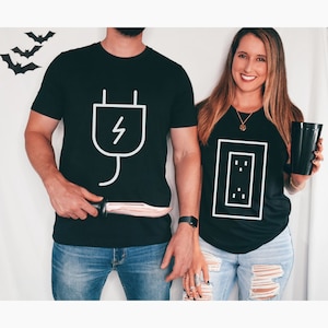 May include: Black t-shirts with white graphics. One shirt features a plug design with a lightning bolt, the other a wall outlet. The couple is wearing blue jeans. Halloween theme.