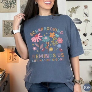 May include: A denim blue t-shirt featuring a colorful floral graphic with the text "SCRAPBOOKING REMINDS US LIFE HAS BEEN GOOD" in a multi-colored font. The design includes various stylized flowers in shades of pink, orange, and blue.