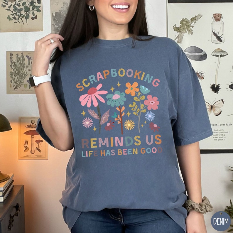 May include: A denim blue t-shirt featuring a colorful floral graphic with the text "SCRAPBOOKING REMINDS US LIFE HAS BEEN GOOD" in a multi-colored font. The design includes various stylized flowers in shades of pink, orange, and blue.