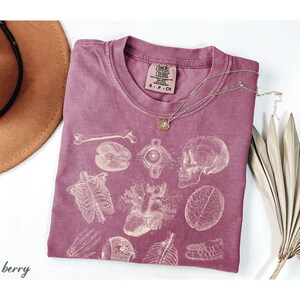 May include: Berry-coloured Comfort Colors t-shirt with a vintage anatomical print of bones, organs, and a skull. A gold necklace with a small pendant rests on the shirt. The shirt is folded on a white surface.