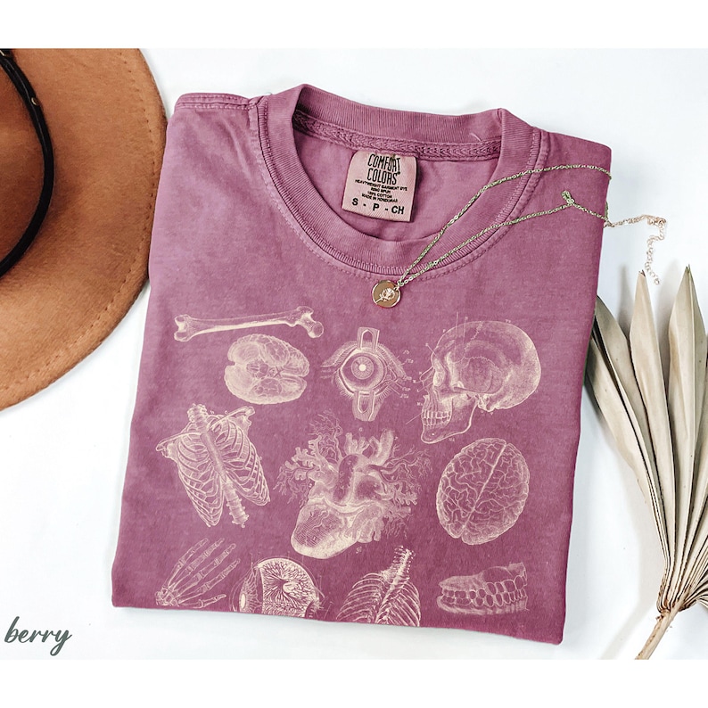 May include: Berry-coloured Comfort Colors t-shirt with a vintage anatomical print of bones, organs, and a skull. A gold necklace with a small pendant rests on the shirt. The shirt is folded on a white surface.