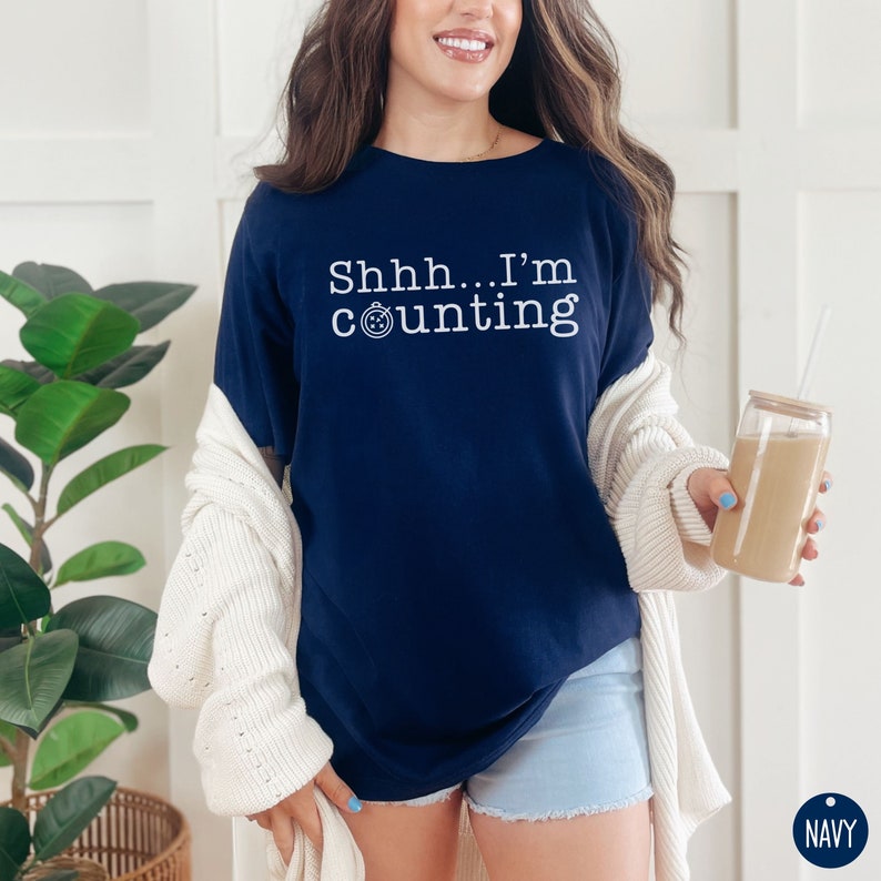 May include: A navy blue t-shirt with the text "Shhh... I'm counting" in white. The shirt features a small clock icon next to the word "counting".
