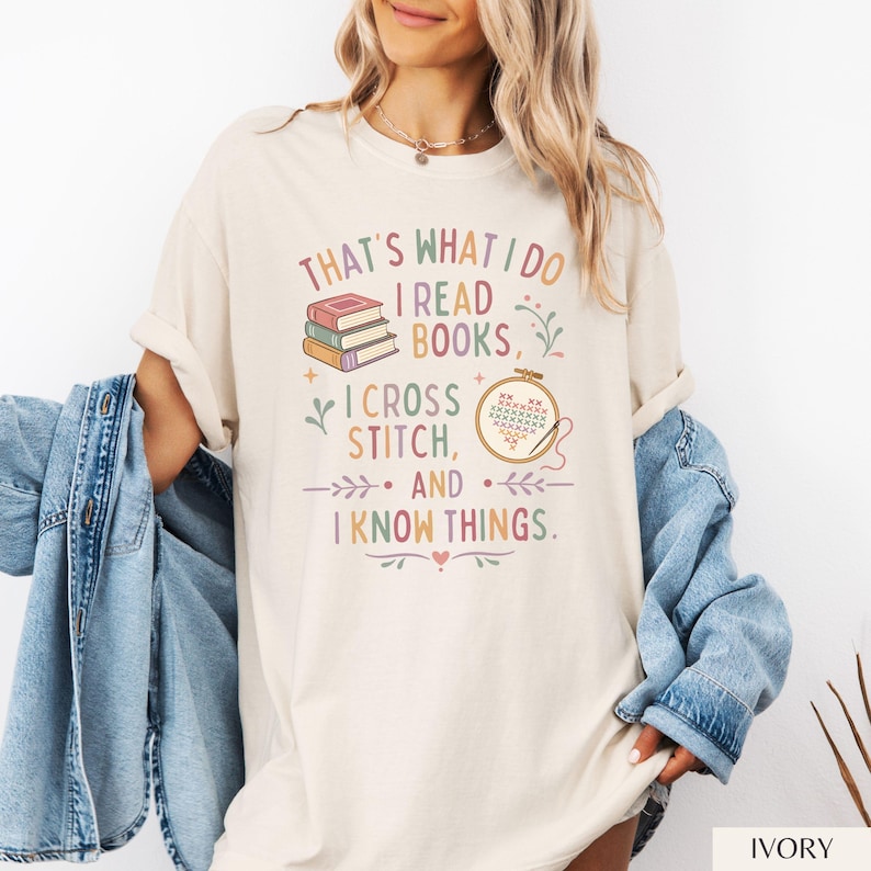 May include: Ivory-colored t-shirt with the text "THAT'S WHAT I DO I READ BOOKS, I CROSS STITCH, AND I KNOW THINGS." The design includes stacked books, a cross-stitch hoop, and decorative floral elements. The shirt is a relaxed fit.