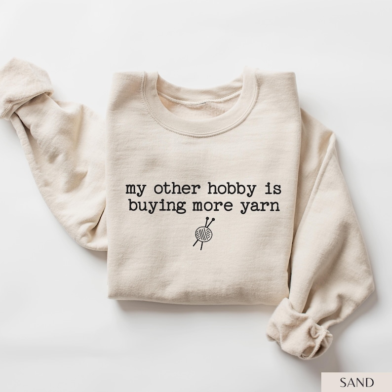 May include: A sand-colored crewneck sweatshirt featuring the black text "my other hobby is buying more yarn" centered on the chest, accompanied by a small graphic illustration of a ball of yarn with two knitting needles crossed through it.