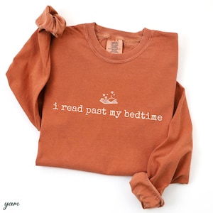 May include: Rust-colored long-sleeve shirt with the text "i read past my bedtime" in white. The shirt features a graphic of an open book with stars above it. The shirt is made of a soft material.