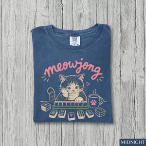 May include: A blue t-shirt featuring a graphic of a tabby cat playing mahjong with the text meowjong in pink script. The design includes a steaming mug with a pink paw print, scattered mahjong tiles, and a small heart above the cat's head.