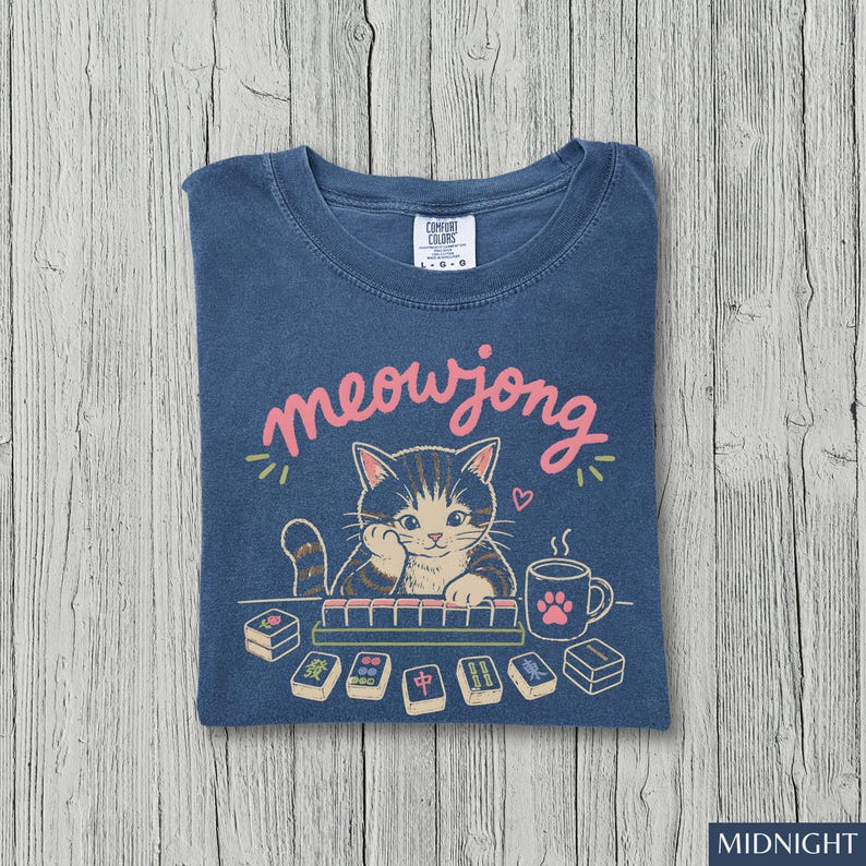 May include: A blue t-shirt featuring a graphic of a tabby cat playing mahjong with the text meowjong in pink script. The design includes a steaming mug with a pink paw print, scattered mahjong tiles, and a small heart above the cat's head.