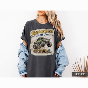 May include: Dark grey t-shirt with a graphic of a monster truck jumping over a car, with the text "Raising Boys Is My Jam" in a vintage style. The shirt is a relaxed fit.
