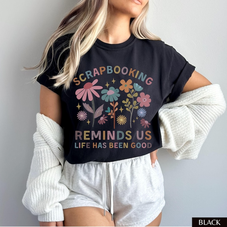 May include: A black short-sleeve t-shirt featuring a colorful floral graphic and the text "SCRAPBOOKING REMINDS US LIFE HAS BEEN GOOD" in a multi-colored font. The design includes various stylized flowers, leaves, and small star accents.