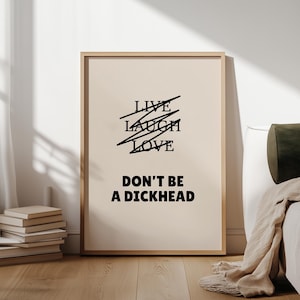 Peut inclure: Une affiche encadrée avec un fond beige et du texte noir. Le texte supérieur indique "LIVE LAUGH LOVE" avec des lignes qui traversent les mots. En dessous, le texte dit "DON'T BE A DICKHEAD". Le cadre est marron clair.