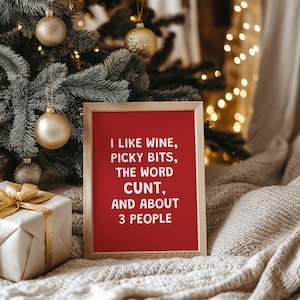 Può includere: Un cartello incorniciato con sfondo rosso e la scritta "I LIKE WINE, PICKY BITS, THE WORD CUNT, AND ABOUT 3 PEOPLE" in bianco. Il cartello è in una cornice di legno, vicino a un albero di Natale con ornamenti dorati e un regalo confezionato.