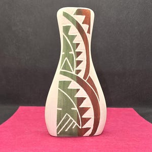 May include: A tall, slender vase with a light beige base color. The surface features a decorative pattern of vertical, curved bands in muted green and dark brown, accented by sharp, triangular cutouts that reveal the light beige color underneath.