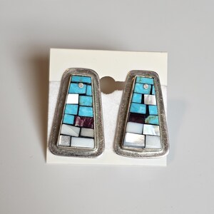 May include: A pair of silver earrings with a geometric design. The earrings are made of turquoise, mother of pearl, and other stones.