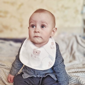 May include: A baby wearing a grey sweater and blue trousers sits on a blanket. The baby is wearing a white bib with a brown teddy bear design.