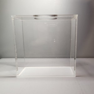 May include: A clear acrylic box with a circular opening in the top. The box is empty and has a square shape.